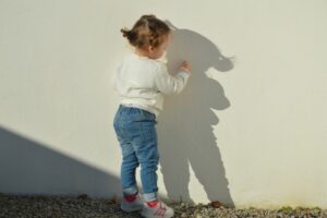 Mädchen im Kleinkindalter steht vor einer weißen Wand und schaut sich seinen Schatten an