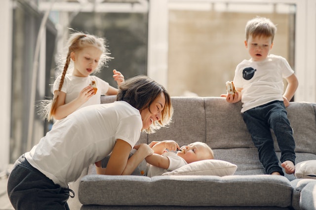 Mutter lacht ihr Baby an, welches auf der Couch liegt. Zwei Kleinkinder stehen daneben und schauen zu.